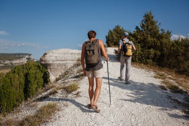 Kayalık yamaçlarda yürüyen iki adam yürüyüş çubuklarını kullanarak sırt çantalarını taşıyor..