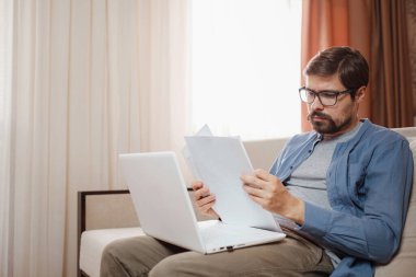 Gözlüklü girişimci bir dizüstü bilgisayarla çalışır ve bir belgeyi ev ofisinde tutar. Elinde kağıt belgeler tutan, iş yerindeki müşterilerle online sohbet eden bir adam..