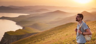 Sırt çantalı genç sporcu kayanın tepesinde duruyor ve yazın günbatımında deniz kıyılarına ve dağlara bakıyor. Açık havada seyahat etmenin yaşam tarzı konsepti.