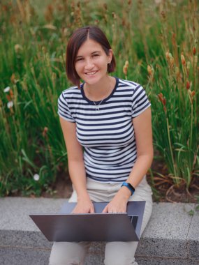 Modern üniversite kampüs parkında dizüstü bilgisayarla çalışan mutlu Asyalı kız öğrenci. Çizgili tişörtlü ve açık renk pantolonlu kadın gri dizüstü bilgisayarda çalışıyor. Öğrenci dışarıda okuyor..