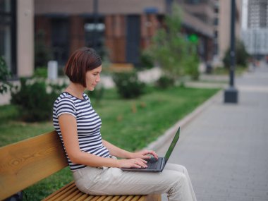 Dizüstü bilgisayarda çalışan öğrenci şehir caddesinde bankta oturuyor..