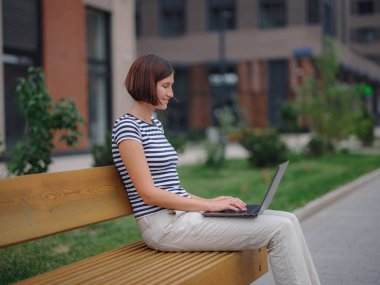 Dizüstü bilgisayarda çalışan öğrenci şehir caddesinde bankta oturuyor..