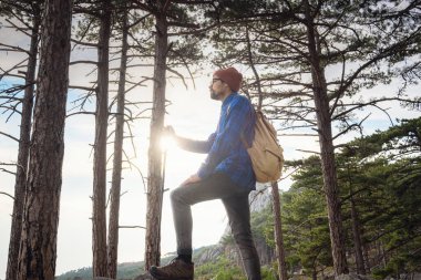 Keşif ve yürüyüş konsepti, doğa ve özgürlük. Genç adam yaz dağlarında bir uçurumun tepesinde çam ormanlarının arasında duruyor ve doğa manzarasının tadını çıkarıyor.
