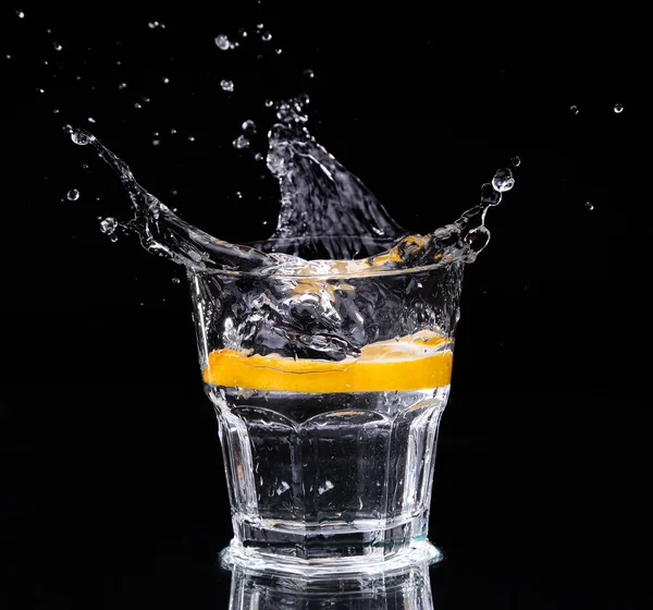 Schijfje citroen spatten in een glas water met een spray van waterdruppels in beweging opgehangen in de lucht boven het glas op een donkere achtergrond. — Stockfoto