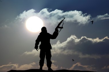 Elinde renkli gökyüzü ve arka planda dağ olan silahlı bir askerin siluet görüntüsü.