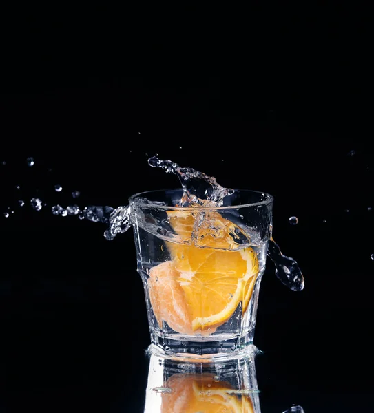 Fetta di limone spruzzata in un bicchiere d'acqua con uno spray di gocce d'acqua in movimento sospese nell'aria sopra il bicchiere su uno sfondo scuro . — Foto Stock