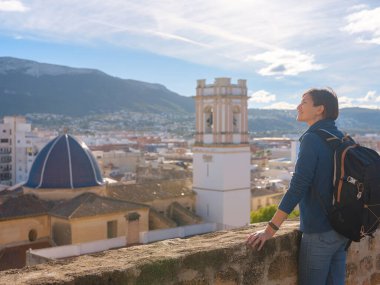 Denia, Alicante, İspanya 'da tarihi şatoyu keşfeden genç bir kadın, aşağıdaki kasabanın manzarası, daha serin mevsim arkaplanlarına karşı. Tarihin ve sükunetin zamansız ortamında harmanlanması.