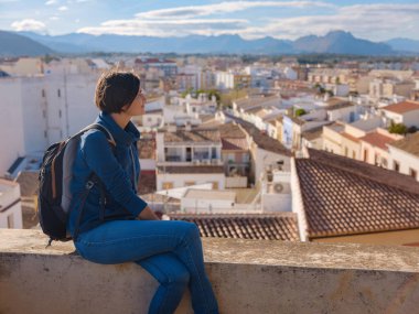 Denia, Alicante, İspanya 'da tarihi şatoyu keşfeden genç bir kadın, aşağıdaki kasabanın manzarası, daha serin mevsim arkaplanlarına karşı. Tarihin ve sükunetin zamansız ortamında harmanlanması.