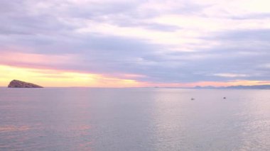 Cala Tio Ximo ve Benidorm adası üzerinde gün batımı manzarası. Sakin kış denizi, yumuşak gün batımı parıltısı, Benidorm yakınlarındaki küçük adanın huzurlu manzarası. Sessiz doğal güzellik, insan yok, sadece deniz ve gökyüzü..