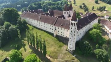 Sankt Paul im Lavanttal 'ın yaz havası manastır kompleksi, çatılar, vadi manzarası, yeşil tepeler, sakin sinema insansız hava aracı hareketi, sıcak atmosfer tonları.