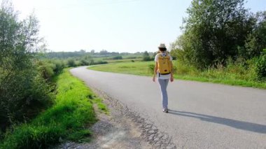 Güneşli bir günde asfalt yolda yürüyen bir kadın yürüyüş macerasına girişiyor. Manzaralı manzaralar ve açık hava ile çevrelenmiş, seyahatin ve açık hava deneyiminin tadını çıkarıyor..