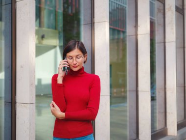 Gözlüklü ve kırmızı balıkçı yakalı bir kadın, Frankfurt 'un merkezinde telefonla konuşuyor, modern binalar ve hızlı şehir hayatı ile çevrili, şehir ortamında profesyonellik ve iletişimi dengeliyor..