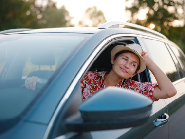 Kırmızı etnik giysili ve şapkalı gezgin kadın yazın Hırvatistan 'ın manzaralı kırsalında araba sürüyor. Etrafı yemyeşil tarlalar ve ormanlarla çevrili, huzurlu ve maceralı bir yolculuktan hoşlanıyor.