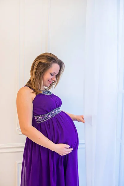 Hermosa mujer embarazada en vestido ultra violeta de pie cerca de la ventana — Foto de Stock