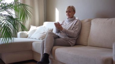 50 yaşında bir kadın kanepeye oturur ve akıllı telefon kullanır.