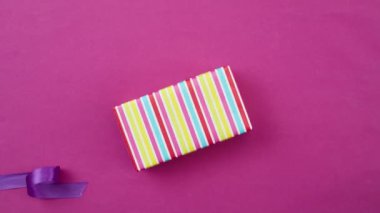 Hareket animasyonunu durdur. Renkli paketlenmiş hediye kutusu ve pembe arka planda mor kurdele, üst görünüm