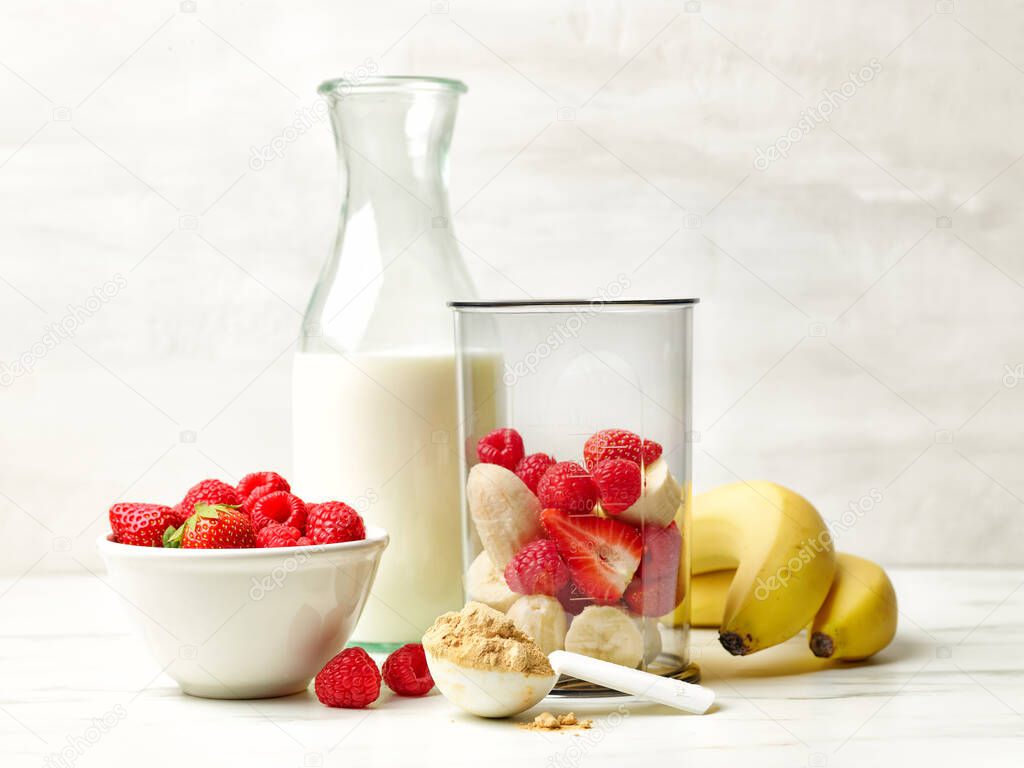 fresh banana pieces and red berries in plastic transparent blender container and milk bottle on kitchen table ready for making healthy breakfast smoothie