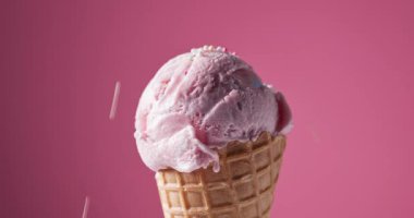 Krep külahında bir kepçe çilekli dondurma pembe arka planda dönüyor, şekerli süslemeler buz topunun üzerine düşüyor. 