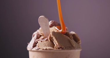 Çikolata ve fıstık dondurması kağıt bardakta dönüyor ve karamel sosu dondurmada akıyor, yemekler mor arka planda kapanıyor.