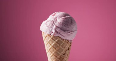 Krep külahında bir kepçe çilekli dondurma pembe arka planda dönüyor, şekerli süslemeler buz topunun üzerine düşüyor. 