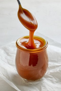 Yeni pişirilmiş ev yapımı karamel sosu beyaz mutfak masasının arka planındaki kaşıktan kavanoza dökülüyor.