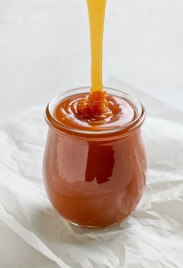 Yeni pişirilmiş ev yapımı karamel sosu beyaz mutfak masasının arka planında kavanoza dökülüyor.