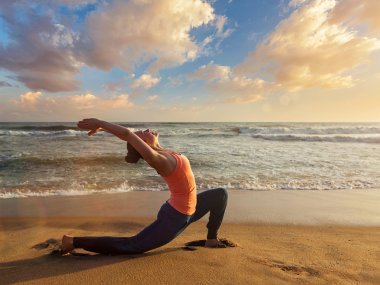 Sportif formda bir kadın gün batımında Anjaneyasana sahilinde yoga yapıyor.