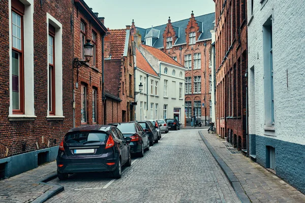 Bruggova ulice s dlážděnou silnicí s zaparkovanými auty a starými středověkými domy. Bruges, Belgie — Stock fotografie