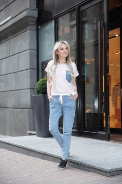 Pantolon ve tişört giymiş genç ve güzel bir bayan dükkanın yanında poz veriyor.