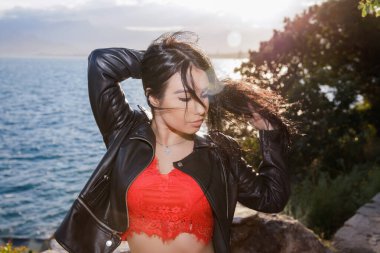 Kırmızı bluz ve ceket giymiş, denizin arka planında poz veren genç bir esmer.