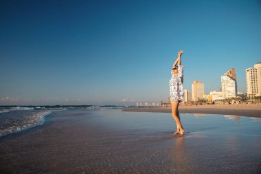 Sahilde poz veren güzel bir kadın. Suda duruyor. Üzerinde tunik ve gözlük var.