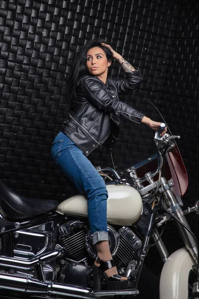 Alluring young lady dressed in a leather jacket and jeans posing on a motorcycle