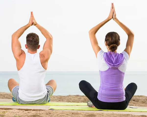 Pareja con piernas cruzadas practica yoga en la playa por la mañana —  Fotos de Stock