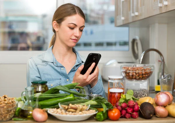 Portrait of glad woman looking at mobile phone while making dish on kitchen at home — Stock Photo, Image
