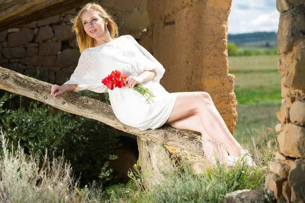 Beyaz elbiseli genç bir kadın elinde gelincik çiçekleriyle ağaç gövdesinde poz veriyor. — Stok fotoğraf
