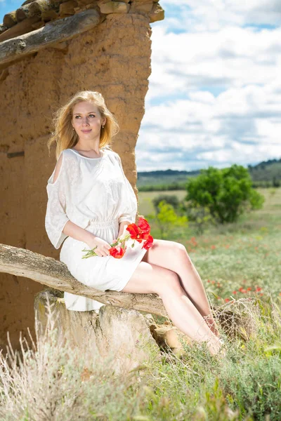 Mladá žena v bílých šatech sedí na stromě a drží mák rostliny — Stock fotografie