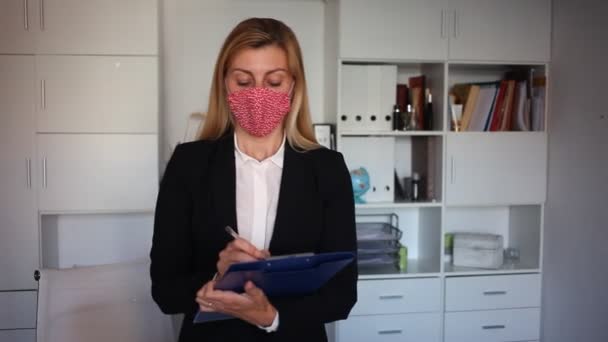 Yüzü maskeli genç bir iş kadını elinde bir panoyla modern şirket ofisine hoş geldin diyor. — Stok video