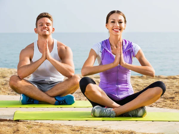 Mulher e homem sentados de pernas cruzadas fazem poses de ioga na praia — Fotografia de Stock