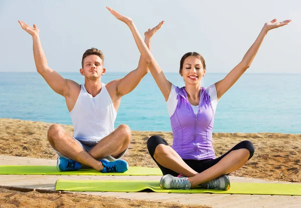 Mulher e homem sentados de pernas cruzadas fazem poses de ioga na praia — Fotografia de Stock