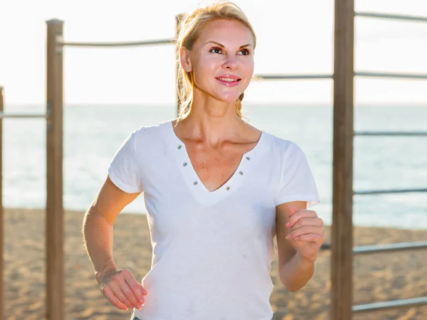 Femme adulte souriante en T-shirt blanc fait du jogging sur la plage — Photo