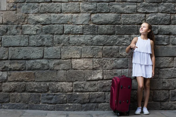 little girl traveler with suitcase leaning against stone wall