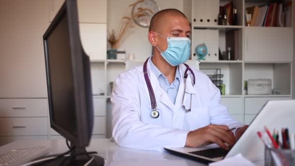 Orvos védő orvosi maszkban ül a munkahelyén számítógéppel az irodájában — Stock videók
