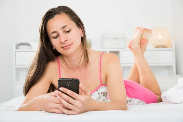 Mujer adulta positiva usando su teléfono mientras está acostada en la cama — Foto de Stock