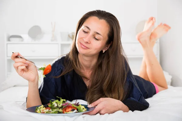 Стройная женщина ест салат в постели дома и улыбается — стоковое фото
