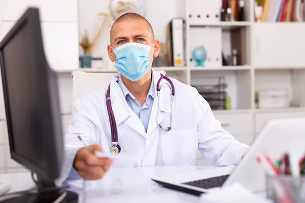 Tıbbi maskeli adam klinikte dizüstü bilgisayarda çalışıyor. — Stok fotoğraf