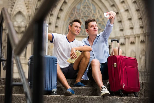 Sorridente coppia maschile con bagagli facendo selfie e seduta — Foto Stock