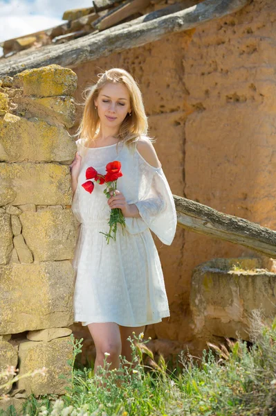 Kobieta w białej sukience stoi w pobliżu kamiennej ściany i gospodarstwa maki roślin — Zdjęcie stockowe