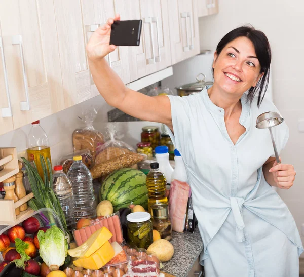 Весела жінка приймає селфі по телефону вчасно готуючи на кухні — стокове фото