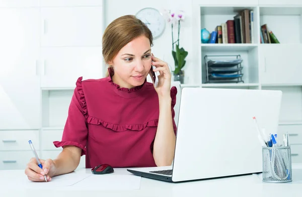 Friendly female employee answering on phone and taking notes — Stock Photo, Image