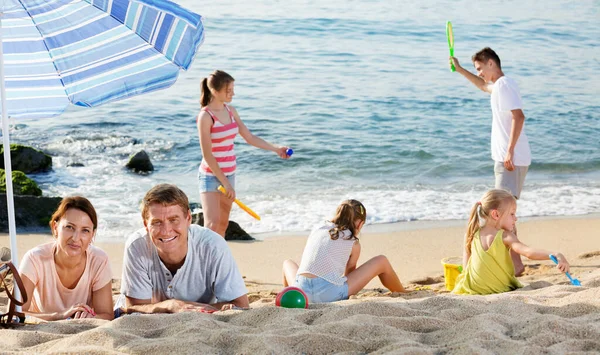 Couple relaxing on beach while their kids playing active games — Stock Photo, Image